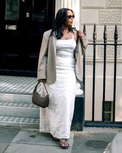 British fashion influencer Karina Marriott poses with oval sunglasses, a tan blazer, white sleeveless linen maxi dress, woven shoulder bag, and heeled flip-flop sandals