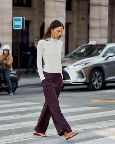 fashion influencer Tylynn Nguyen walking across a NYC crosswalk wearing a white ribbed long-sleeve top, eggplant colored trousers, and red and black flat flip-flop sandals.