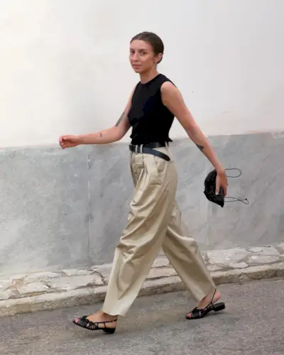 British influencer Brittany Bathgate walking on a street in Molico, Spain wearing a black crewneck tank top, black leather belt, minim black pouch clutch bag, pleated khaki pants, and black woven slingback sandals