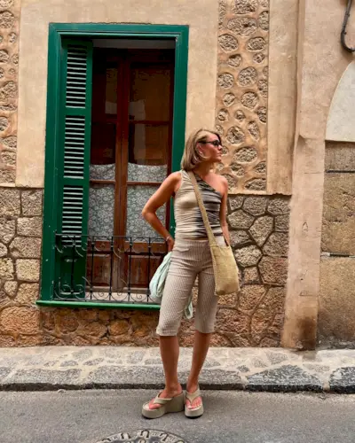 Influencer wears platform flip-flops with capri pants and a one-shoulder top. She's styling a raffia cross-body bag over her shoulders and is wearing dark sunglasses.