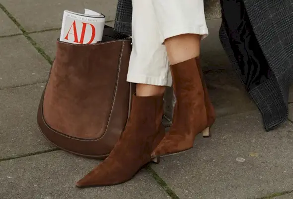 Woman sitting on bench wearing brown suede boots with gray coat and white jeans.