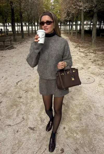 Black turtleneck outfit is shown in a photo of a French woman in the park wearing a brown stretchy headband with brown sunglasses and a black turtleneck layered under a gray crewneck mohair sweater styled with a matching mini shirt, black sheer tights, brown loafers, and a brown Hermes bag