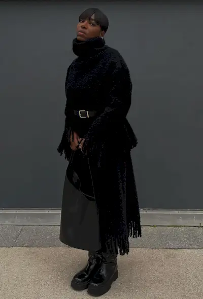Black turtleneck outfit is shown in a photo of a French woman inside wearing a chunky black turtleneck sweater with a black belt ontop styled with a black knit fringe skirt, black tote bag, and black knee-high flat boots
