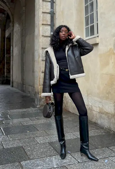 A black turtleneck outfit is shown in a photo of a French woman on the sidewalk wearing a black shearling leather jacket over a black turtleneck with a black mini skirt, designer belt, black tights, black knee-high boots, and a small bag