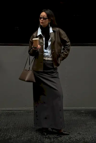 A black turtleneck outfit is shown in a photo of a woman standing on the sidewalk wearing a brown bomber jacket layered over a white button-down shirt over a black turtleneck sweater styled with a white belt, gray maxi skirt, ankle boots, and a brown shoulder bag