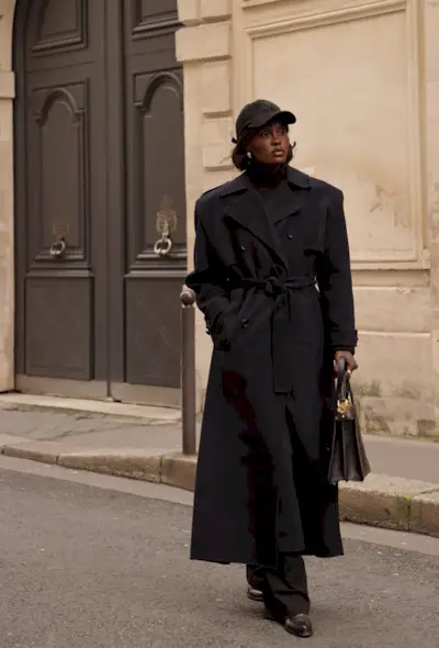 A black turtleneck outfit is shown in a photo of a woman standing on the sidewalk wearing a black baseball cap with a black turtleneck, navy trench coat, black trousers, black loafers, and a black bag