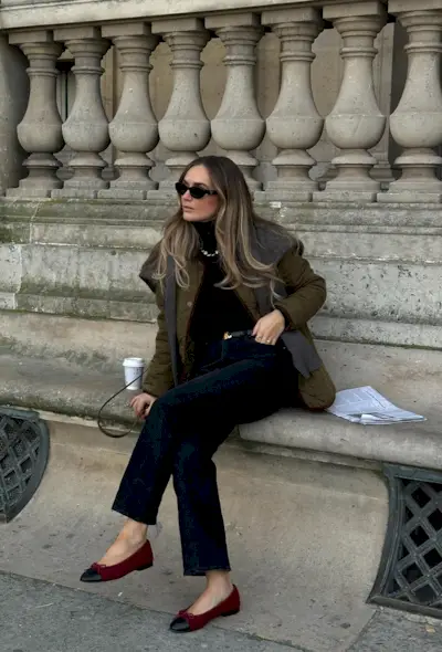 A black turtleneck outfit is shown in a photo of a woman sitting on a park bench wearing a green quilted jacket with a gray cardigan layered over it styled with a black turtleneck, black belt, skinny jeans, and red ballet flats