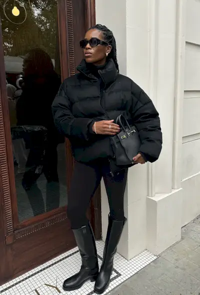 A black turtleneck outfit is shown in a photo of a woman standing on the sidewalk wearing black sunglasses with a cropped black puffer coat layered over a black turtleneck styled with black leggings, black knee-high riding boots, and a black handbag