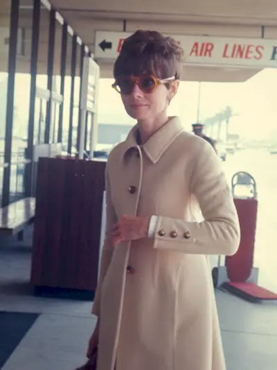 Audrey Hepburn arrives at the airport wearing a cream coat and round acetate sunglasses