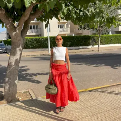 Fashion person Evie Mills has a red skirt outfit. She pairs her red skirt with a white cropped top, khaki green dad sandals and khaki raffia bag and orange ombree sunglasses. She is stood smiling at the camera in recent image taken from her Instagram.