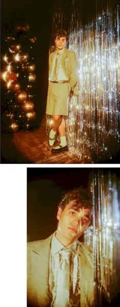 David Iacono stands in front of a silver tinsel wall wearing a cream shorts suit by Wolk Morais with black and white penny loafers paired with white socks.