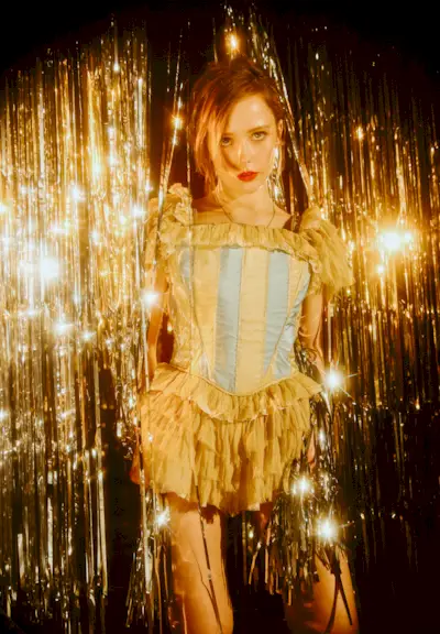 Suzanna Son from Fear Street: Prom Queen is standing in a silver tinsel backdrop wearing a Gabe Gordon minidress with ruffled skirt and gold and blue striped bustier top.