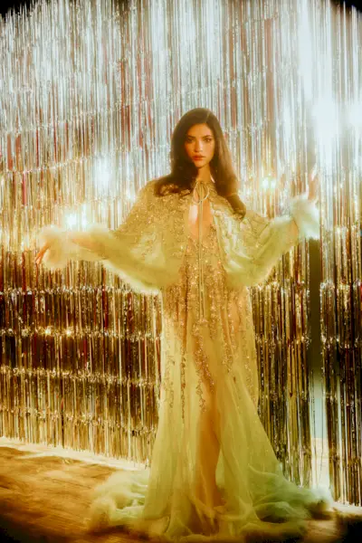Ella Rubin stands in front of a silver tinsel backdrop wearing a floor-length mint green Valentino gown with jewel embellishments and furry detailing at the hemline and a matching capelet.
