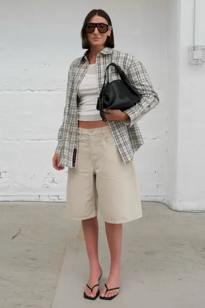 A woman wearing a plaid shirt, white tank top, tan shorts, and black thong sandals