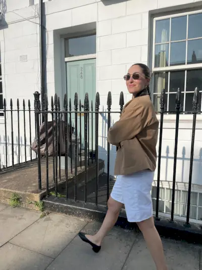 A woman wearing a barn jacket with long white shorts and pumps.