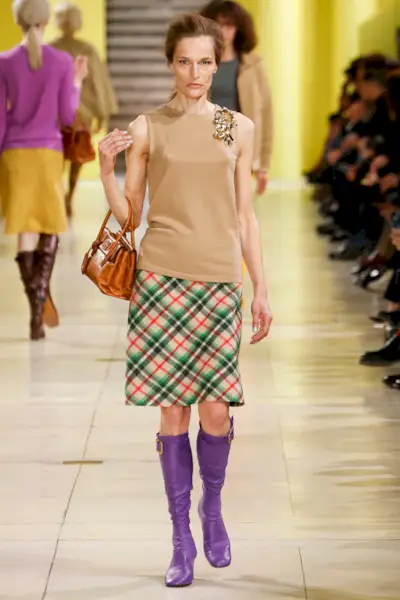 A Miu Miu model wearing purple leather boots, a plaid skirt, and tan tank top