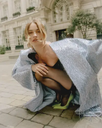Photo of Lily James for Mommy's Reviews's August cover story. She is kneeling down outside of a hotel in London wearing a sky blue overcoat with fringe detailing, a black dress, and green pumps.