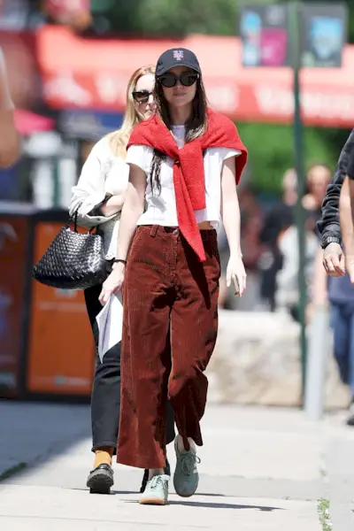 Dakota Johnson wears a baseball cap, black sunglasses, a white t-shirt, corduroy pants, sneakers, and a red sweater tied around her neck.