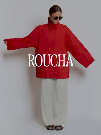 A photo of a model wearing a Roucha red sporty jacket and cream pants. Text says 'Roucha in white capital serif letters.