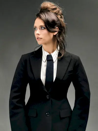 Mia Tomlinson looking off camera. She is wearing a black blazer with exaggerated hips, a white pinstripe shirt, and black tie.