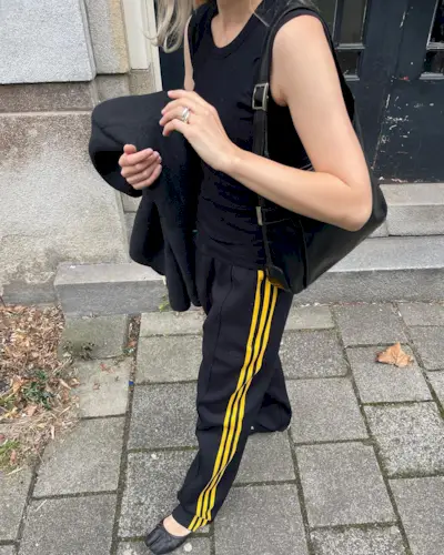 Dutch style influencer Stephanie Broek walking on the sidewalk wearing a black tank top, black Adidas track pants with yellow stripes, and black Margiela ballet flats