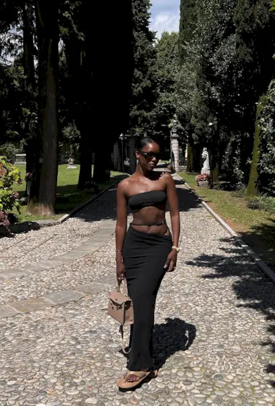 a black maxi skirt outfit showcasing a woman wearing black sunglasses with a black bandeau bikini top and a low-slung black maxi skirt with a tan hermes bag and tan flip flops