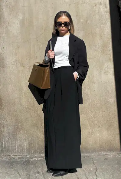 a black maxi skirt outfit showcasing a woman wearing a black headband with black sunglasses, silver hoops, a black blazer, t-shirt, black long skirt, black ankle boots, and a tan handbag