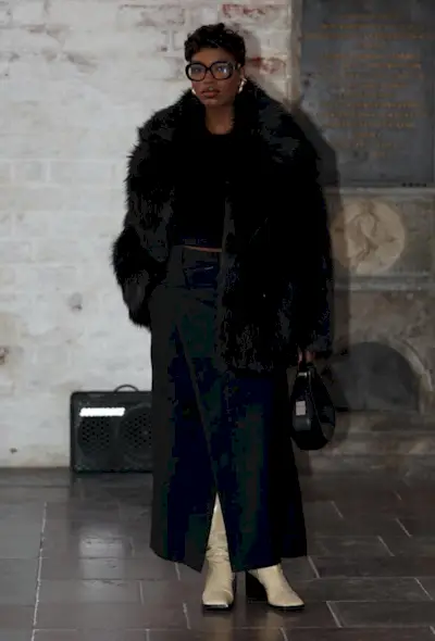 a black skirt maxi outfit showcasing a woman wearing black glasses with gold earrings, a black fur coat over a cropped black sweater and a black maxi skirt and white knee-high boots