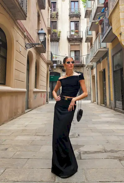 a black maxi skirt outfit shown on a woman wearing gold sunglasses, gold earrings, a black off-the-shoulder top, a black and gold belt, a black heart-shaped bag, and a black column maxi skirt