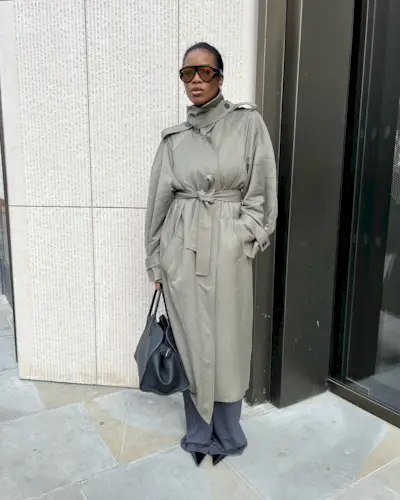 A woman wears a trench coat, with gray trousers, black pointed toe heels, and a black tote bag.