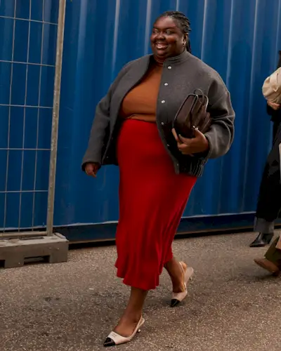 Abi wears a gray bomber jacket, orange sweater, red slip skirt, and two-tone slingback heels.