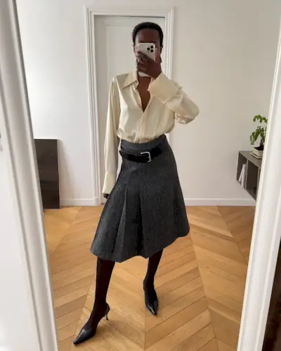 Sylvie wears an ivory silk blouse, gray pleated midi skirt, a black belt, black sheer tights, and black leather pumps.