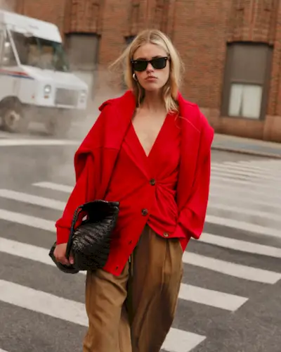 A woman wears a red cardigan, brown trousers, and black bag with a red sweater over her shoulders.
