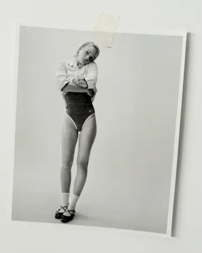 A photo of Esther McGregor taped to a white board. In the photo, she is holding up her white polo sweater to reveal her Celine swimsuit. She is also wearing white socks and strappy flats.