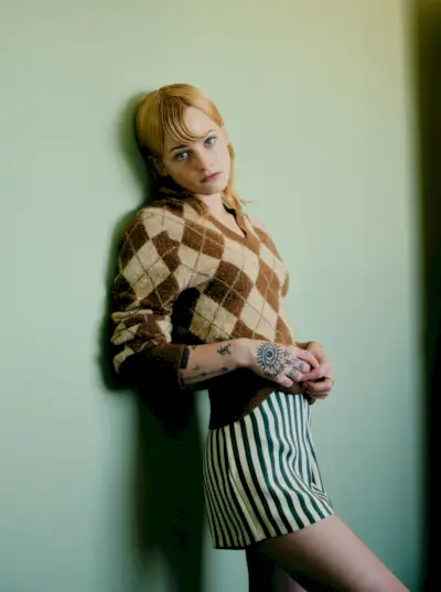 Esther McGregor leans up against a light green wall. She is wearing a brown and tan Coach argyle sweater and Bode blue and white stripe silk shorts.