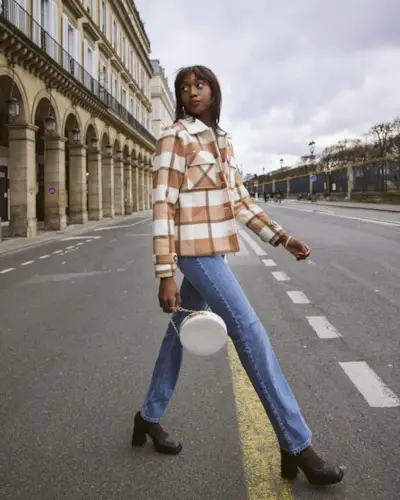 A woman wears a brown and white plaid jacket, blue straight leg jeans, brown pumps, and a white circular bag.