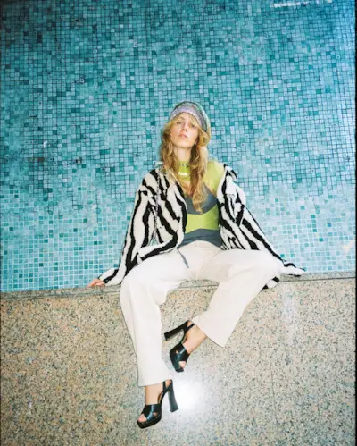 A woman wears a black and white stripe blouse, green top, white jeans, black heels.