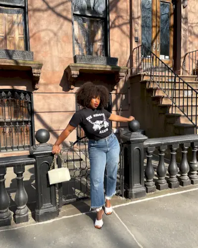 A woman wears a black graphic t-shirt, blue crop jeans, white heels, and a handbag.