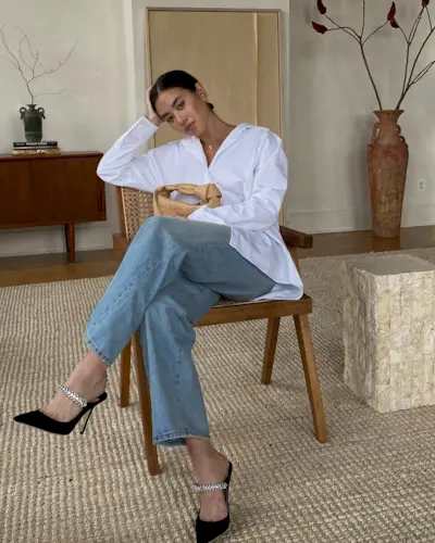 A woman wears a white button-down shirt, blue jeans, and black heels with crystal embellishments.