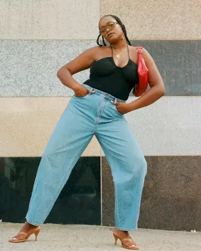 A woman wears a black halterneck top, red bag, blue jeans, and nude heels