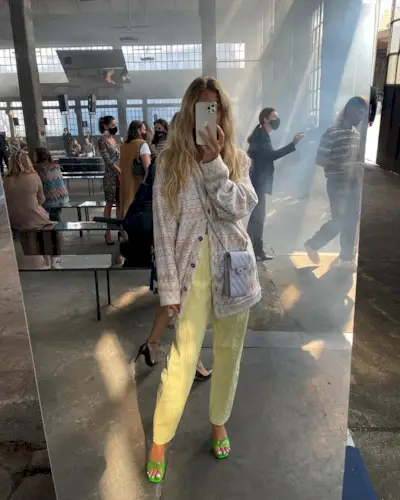 A woman wears yellow trousers, a colorful cardigan, green heels, and a crossbody bag.
