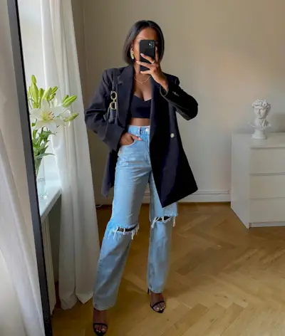 A woman wears a black blazer, black bralette, blue ribbed jeans, black heels, and a black dior bag.