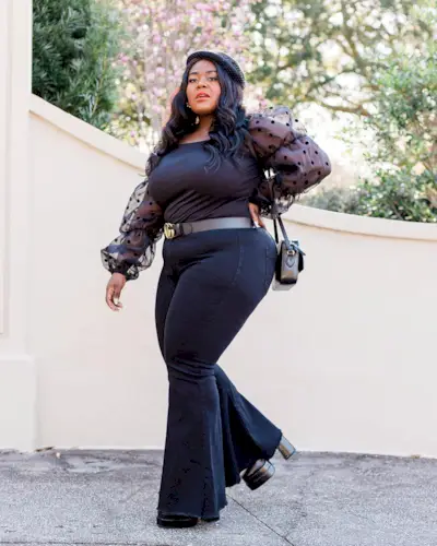 A woman wears a black sheer blouse, gucci belt, black flare jeans, black platform heels, and a black bag.