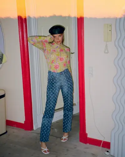 A woman wears a floral button-down shirt, printed blue jeans, white sandals, and a black hat.