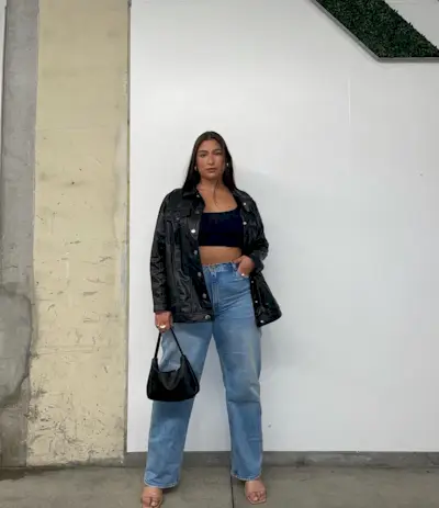 A woman wears an oversize leather jacket, black cropped top, blue jeans, a black bag, and nude mules.