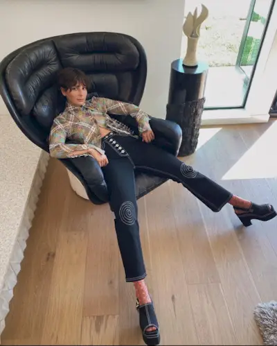 A woman wears a plaid top, blue jeans with swirls, and black clogs.