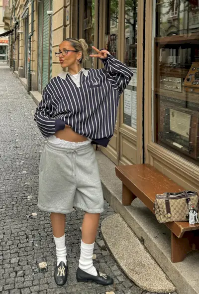 A woman wearing striped rugby polo with gray sweatshorts, white socks, and black loafers.