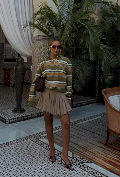 A woman wearing a striped green and brown polo shirt with a pleated tan miniskirt, brown mules, and a brown shoulder bag.