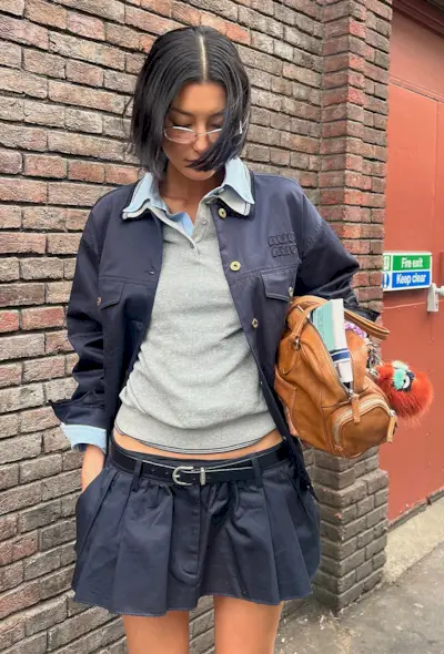 A woman wearing a blue jacket over a blue button-down shirt and gray polo styled with a navy pleated miniskirt and brown bag.
