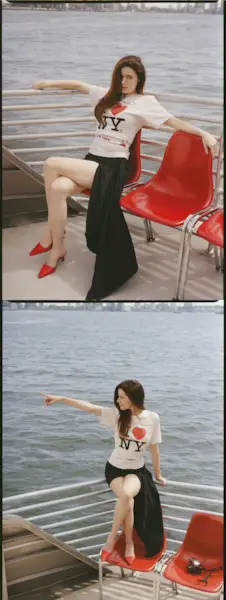 Molly Gordon sitting outside on a ferry boat. She is seated in a red plastic chair, her arms perched on the railing behind her. She is wearing an 'I Love NY T-shirt with a black skirt and red heels.
